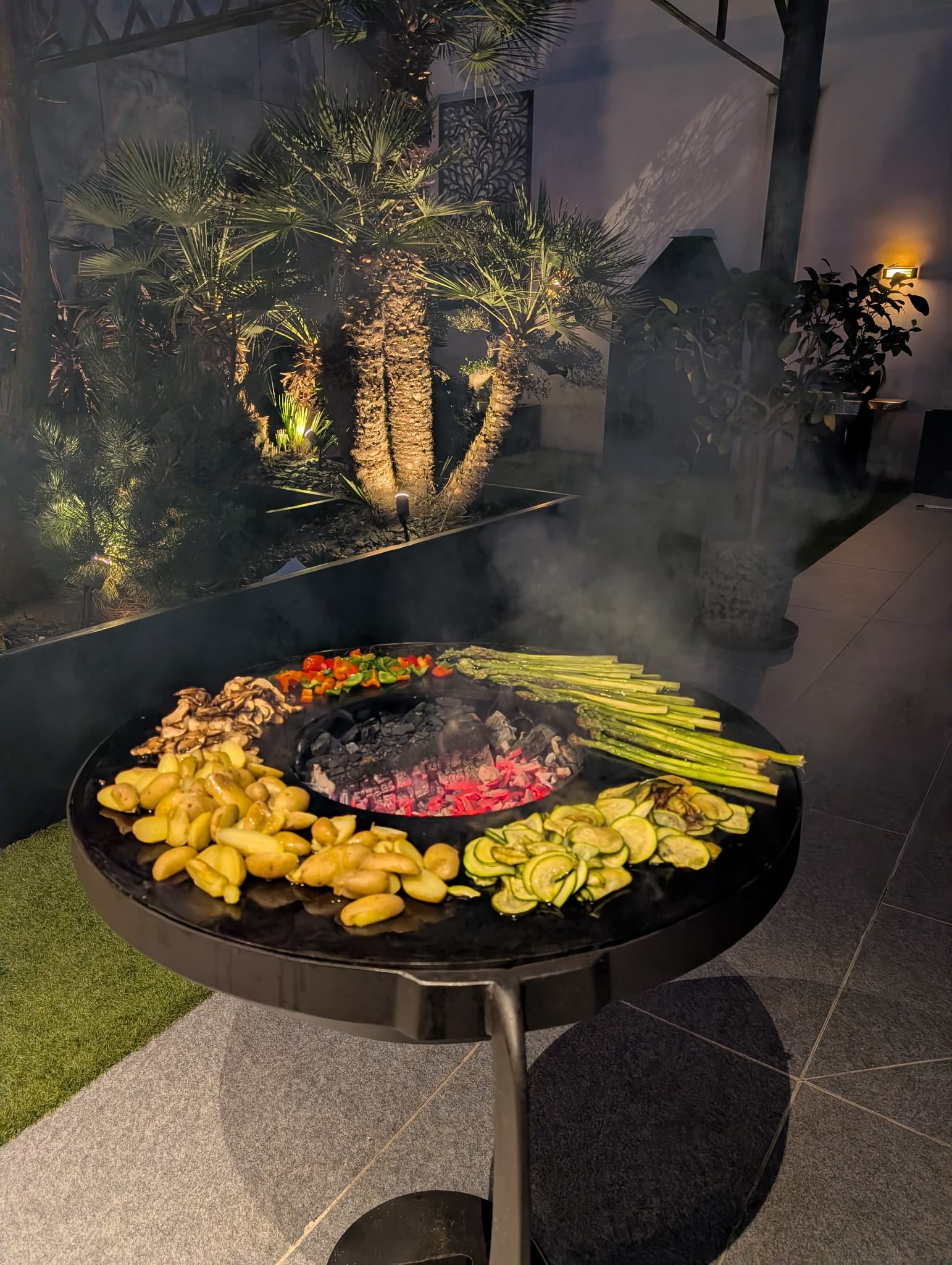 Brasero corten en cuisson sous des palmiers éclairés la nuit