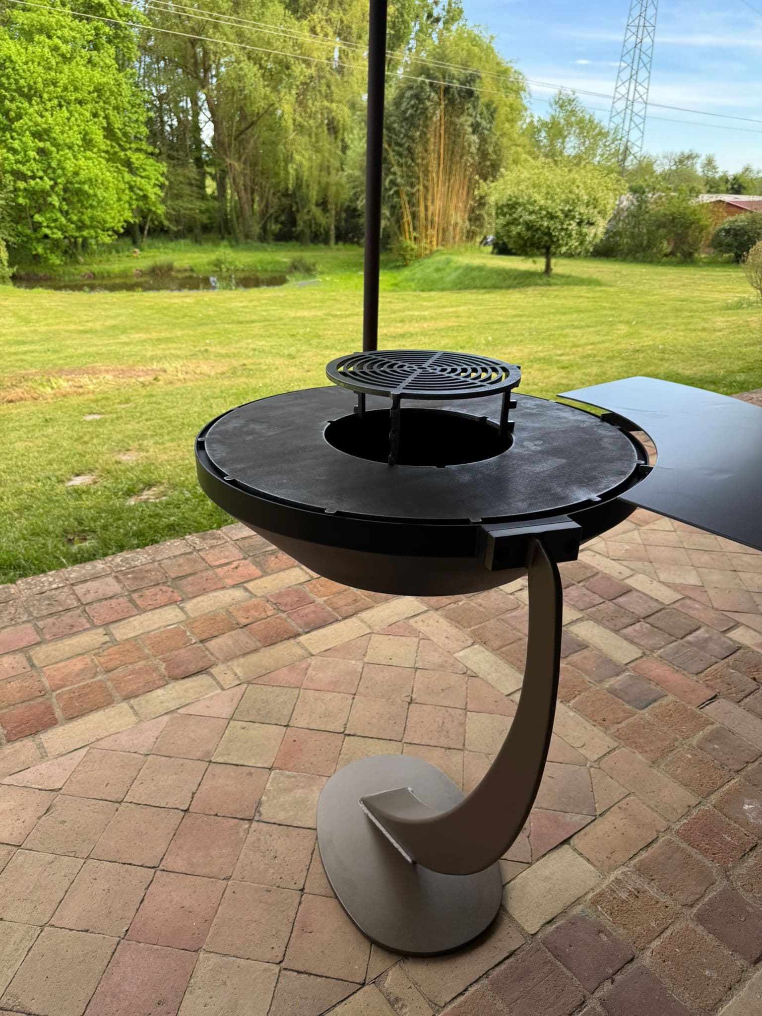 Brasero plancha sur une terrasse en briques, face à un jardin avec étang
