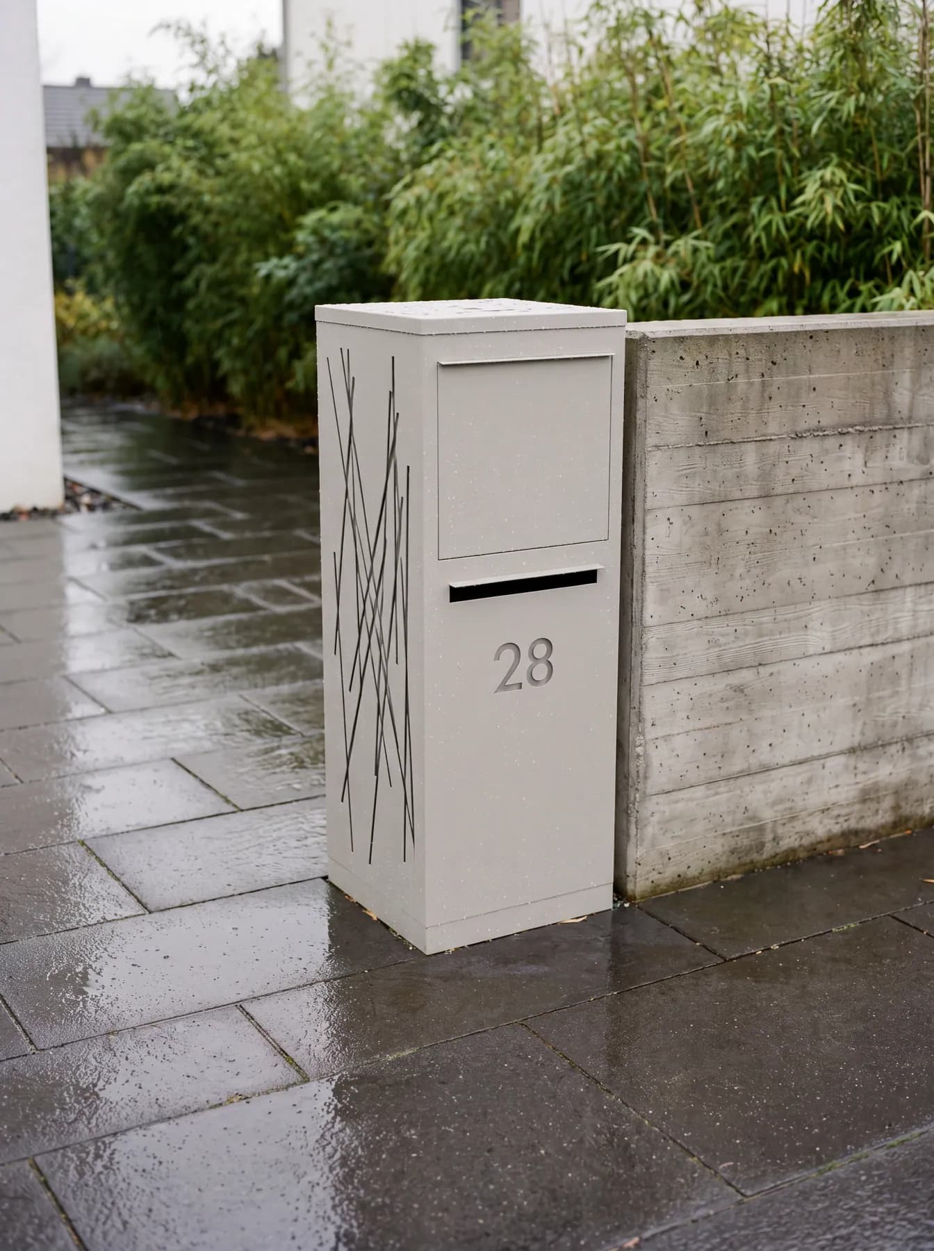 Boîte aux lettres colis gris pierre à motif mikado sous la pluie, devant une haie de bambous